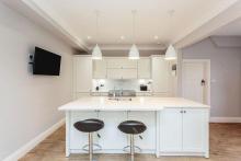 White kitchen with breakfast bar on the island and 2 stools. Behind this are tall housings, wall units and range cooker.